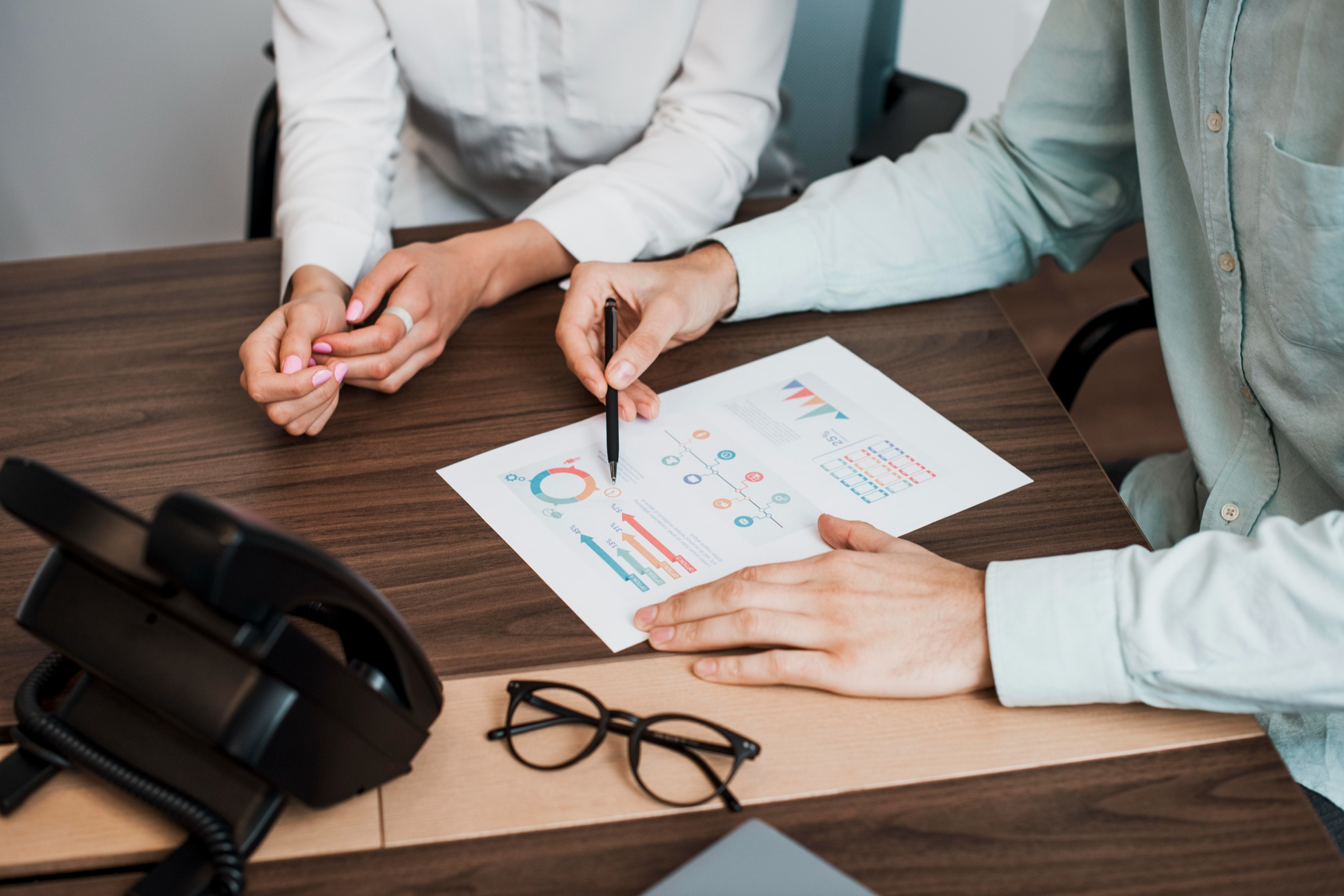 Two people analyzing colorful charts on a desk, with a pen pointing at data and glasses nearby.