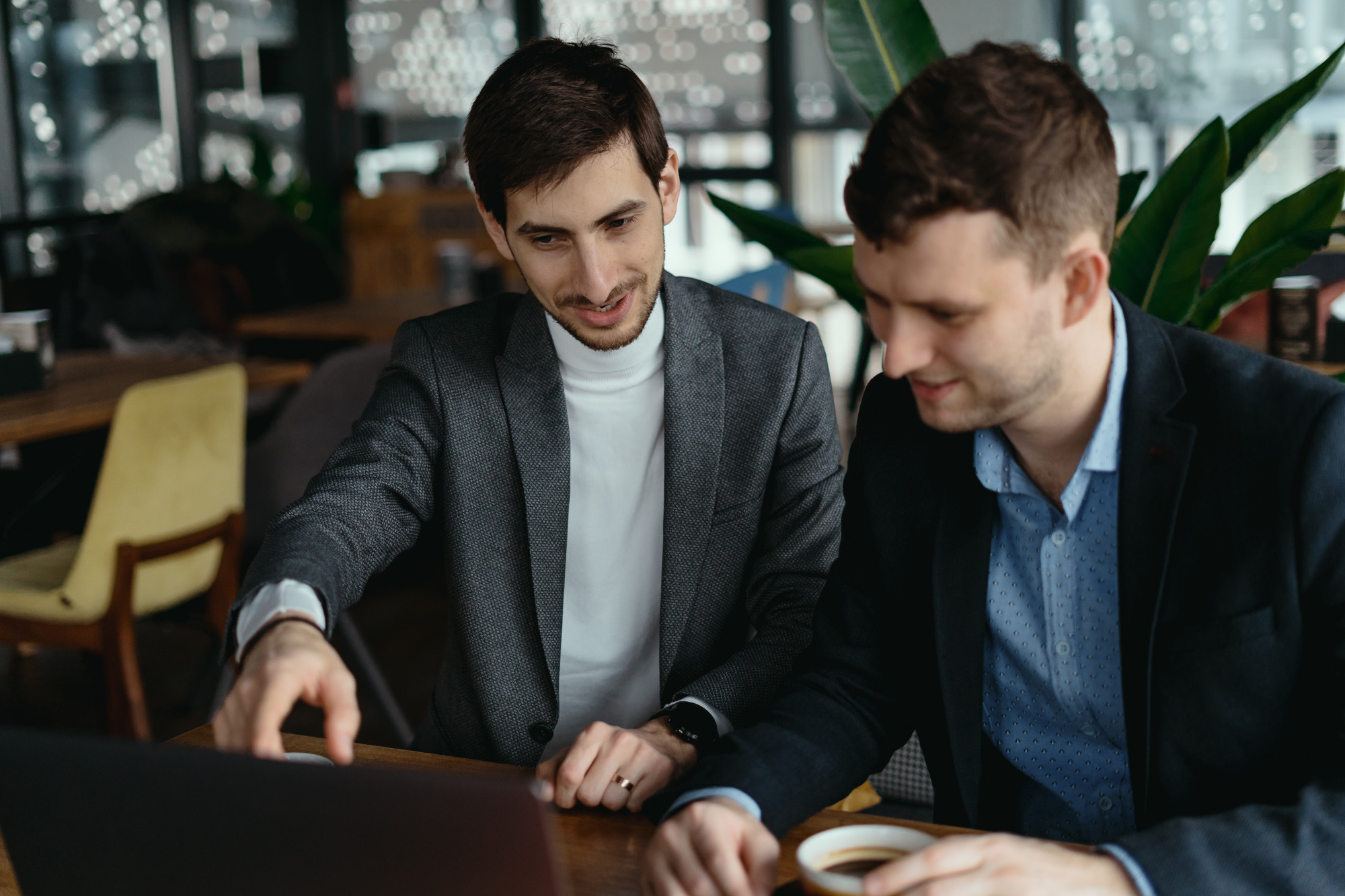 Two men in suits discussing at a table with a laptop and coffee.