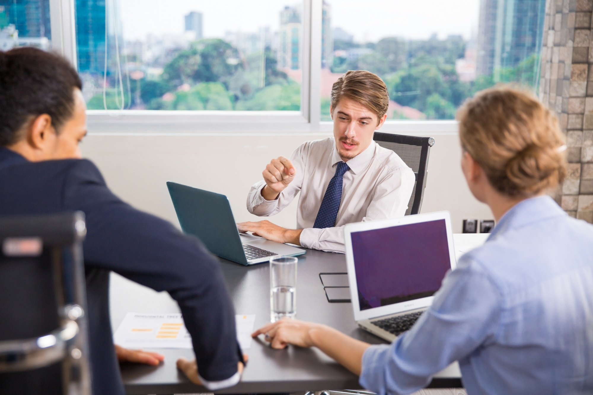 Three people in a meeting with laptops and documents in a bright office.