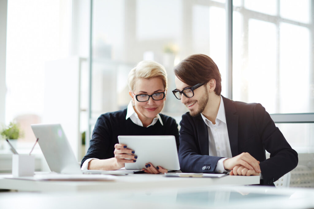 Man and woman in glasses smiling at a tablet in an office.